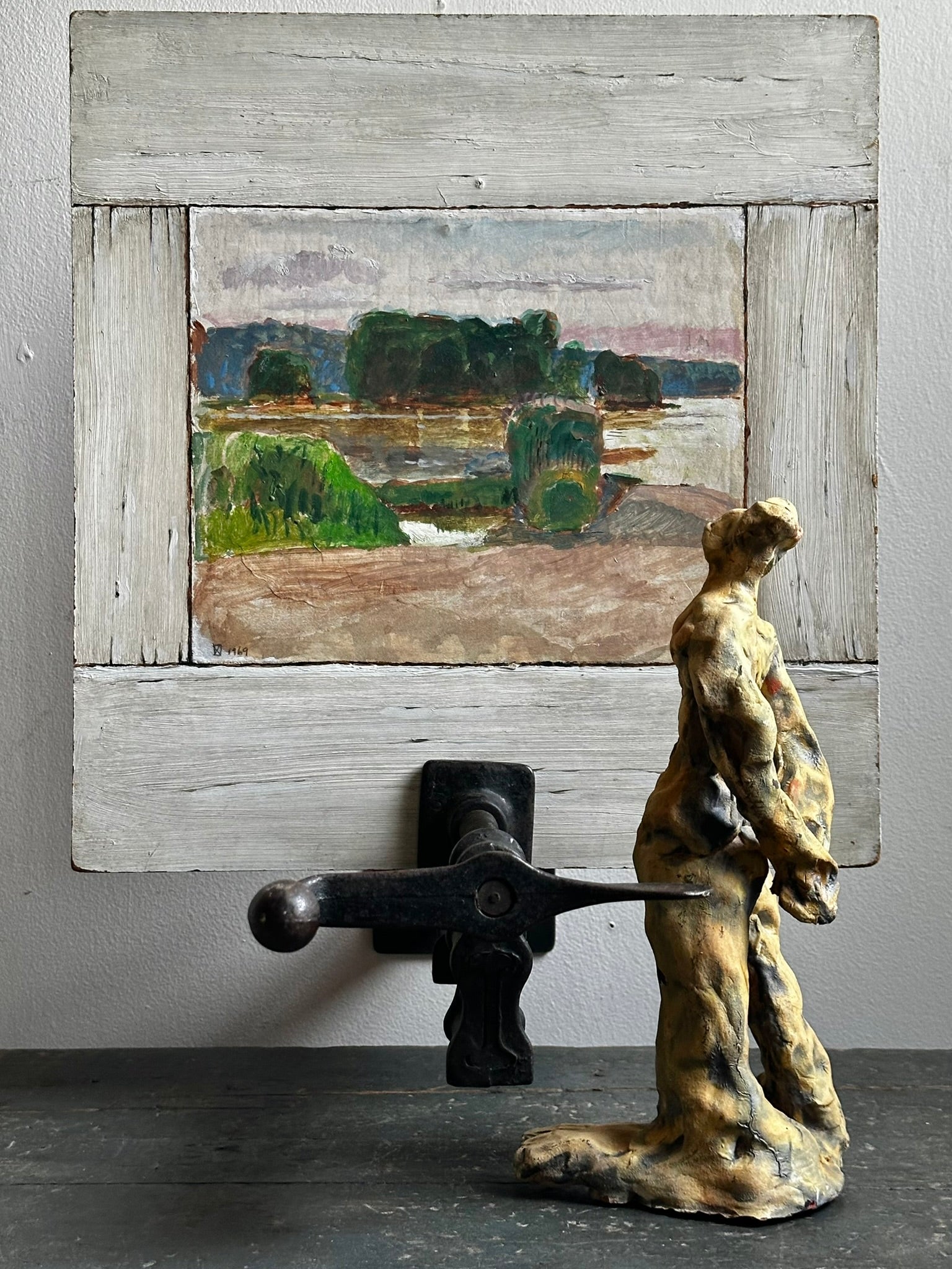 A rustic framed landscape painting displayed on a wall above a sculptural figure and black metal object, with the painting as the likely product shown in a styled interior setting.